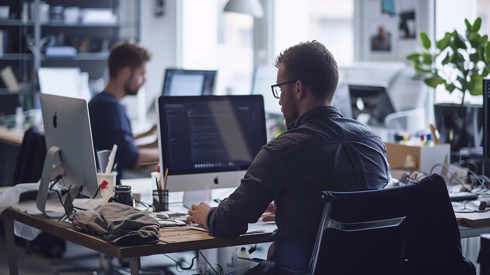 Two developers work at separate desks in a bright office, one facing a large monitor full of code